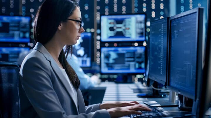 Femme portant des lunettes à un bureau, tapant sur un clavier et regardant deux écrans